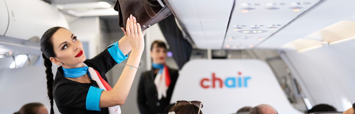 Flight attendant in a black and blue uniform places luggage in an overhead compartment inside a plane cabin. Passengers are seated nearby.